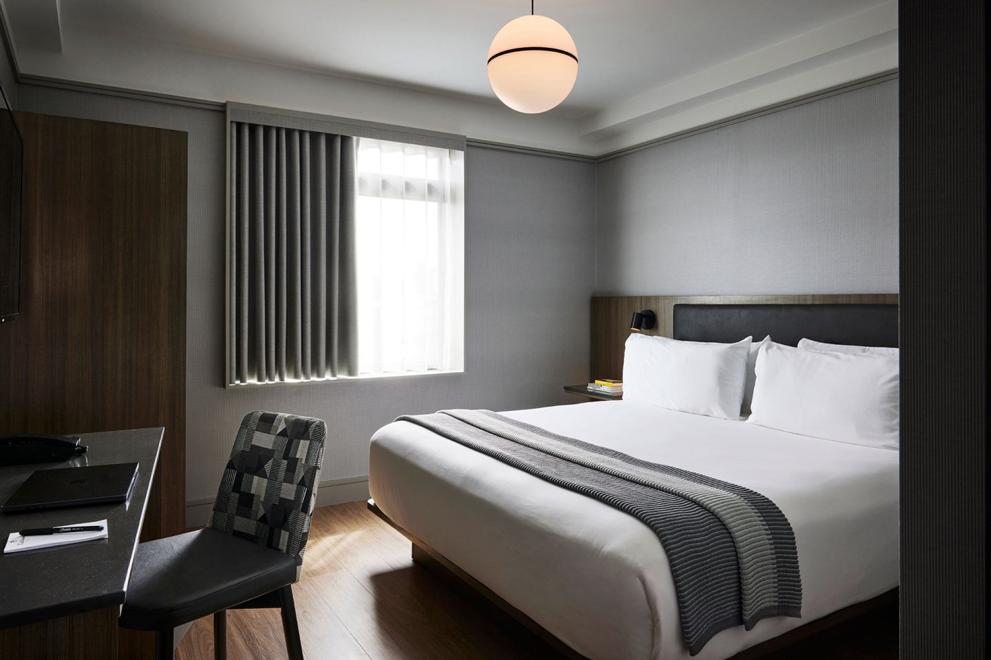 Modern hotel room with a large bed, white linens, and gray-striped blanket. A desk with a chair faces the wall, and a window with gray curtains lets in natural light. Overhead, a round ceiling light is on.