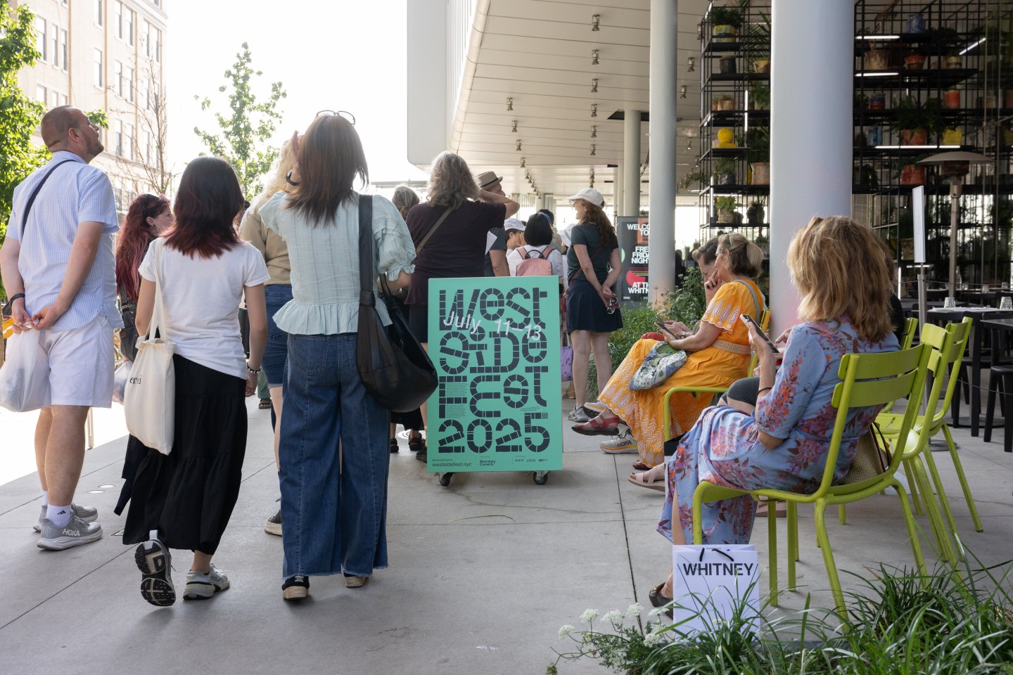 People gather on a sunlit outdoor terrace outside a museum, with a crowd standing and others seated on bright green chairs along a café area. In the center foreground, a teal sign on wheels reads “West Side Fest 2025 – July 11–13.” Behind the crowd, the museum’s modern exterior features tall white columns and glass walls with plants visible inside. A Whitney Museum shopping bag sits near the seated visitors.