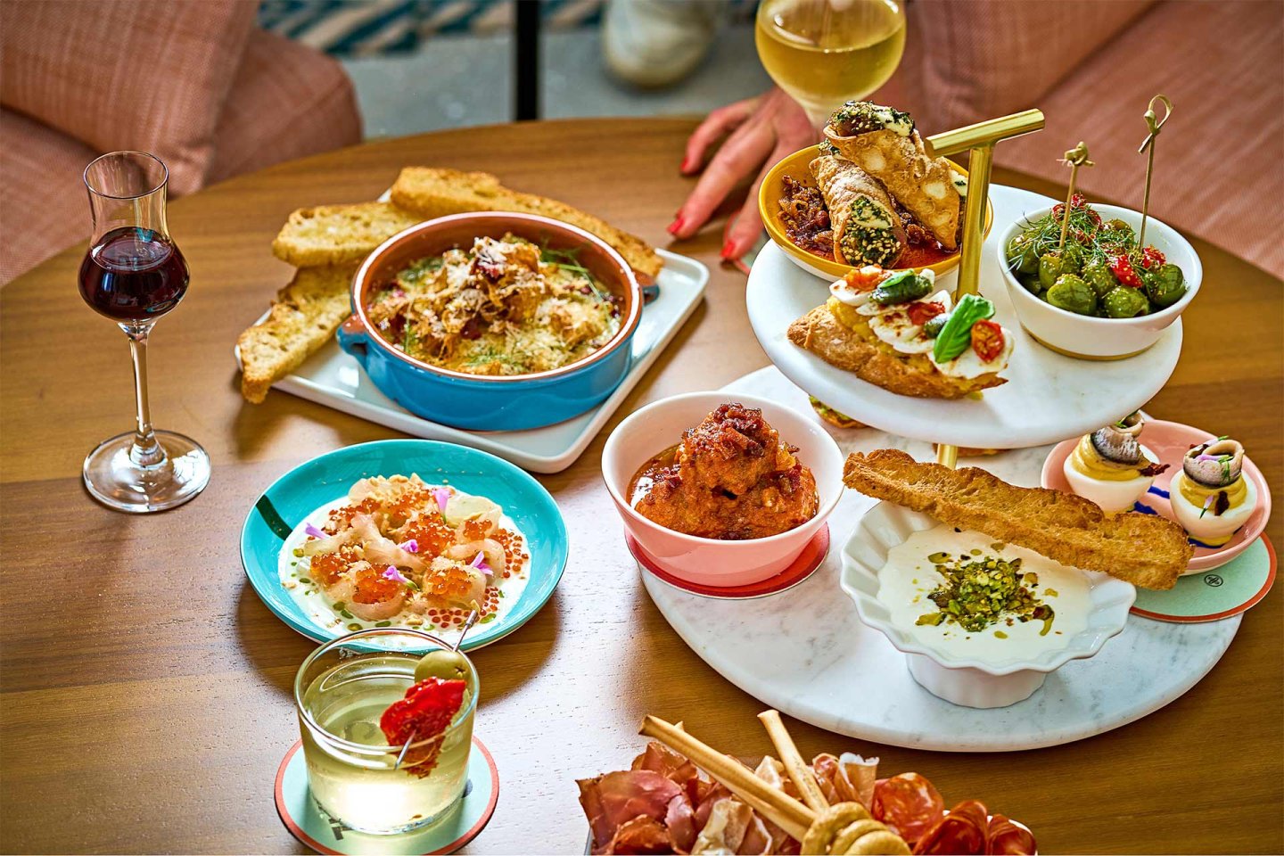 A wooden table set with various appetizers, bread, meatballs, olives, shrimp, and desserts, along with glasses of white and red wine; two people’s hands are partially visible.