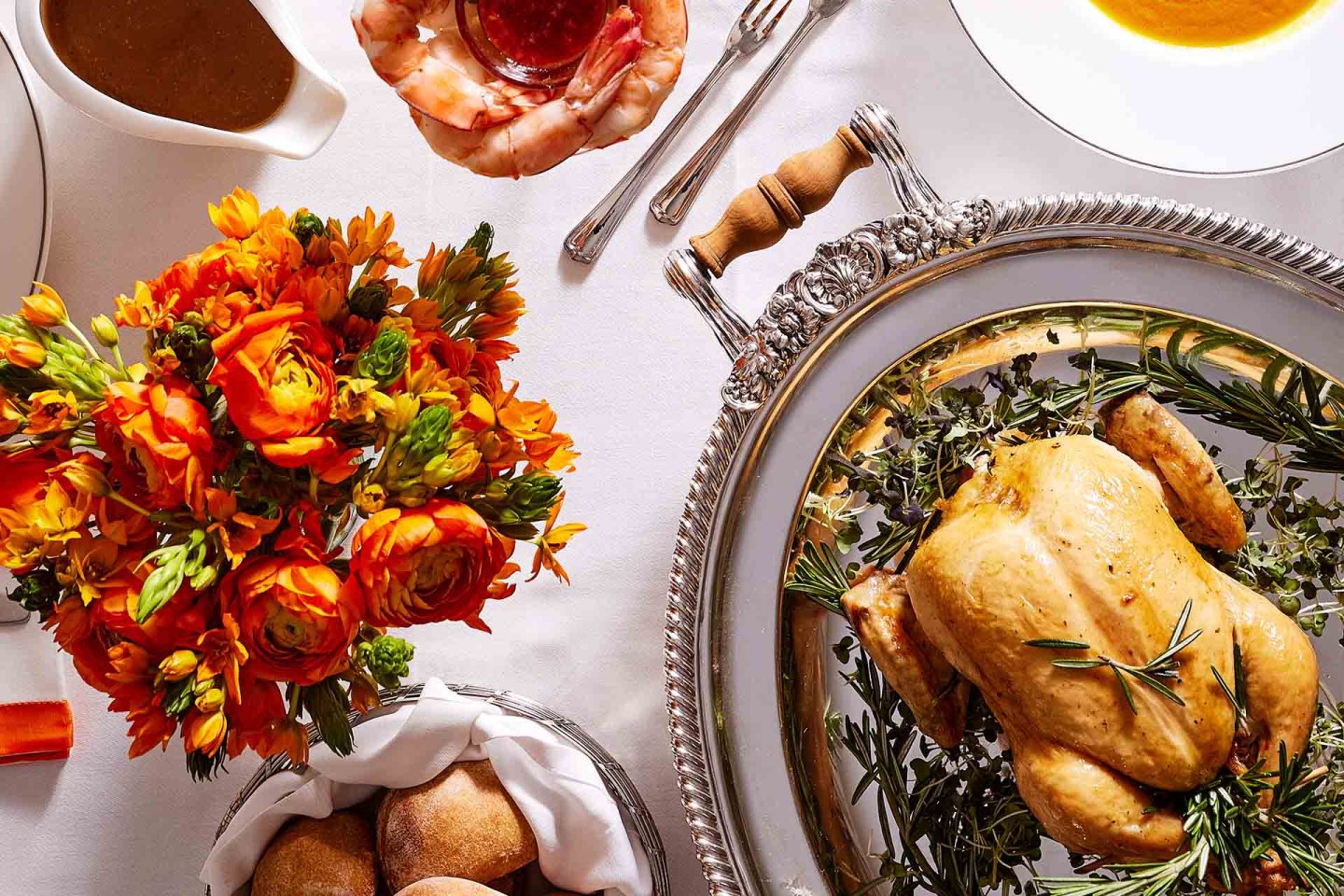 A roast chicken on a silver tray with herbs, orange flowers in a vase, bread rolls, shrimp cocktail, gravy, and soup, all arranged on a white tablecloth.