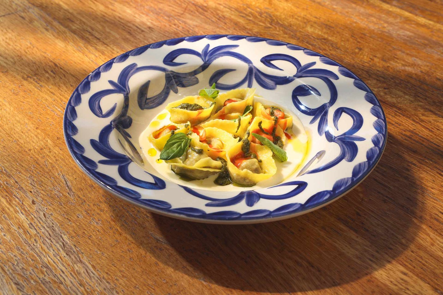 A decorative blue and white plate filled with stuffed pasta shells, garnished with fresh basil leaves and drizzled with olive oil, sits on a wooden table in warm sunlight.