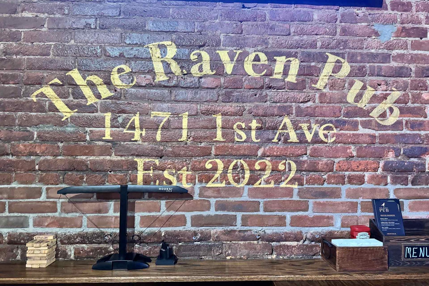 A brick wall displays "The Raven Pub, 1471 1st Ave, Est. 2022" in yellow letters. On the wooden bar below are a scale, Jenga blocks, a menu, and other small items.