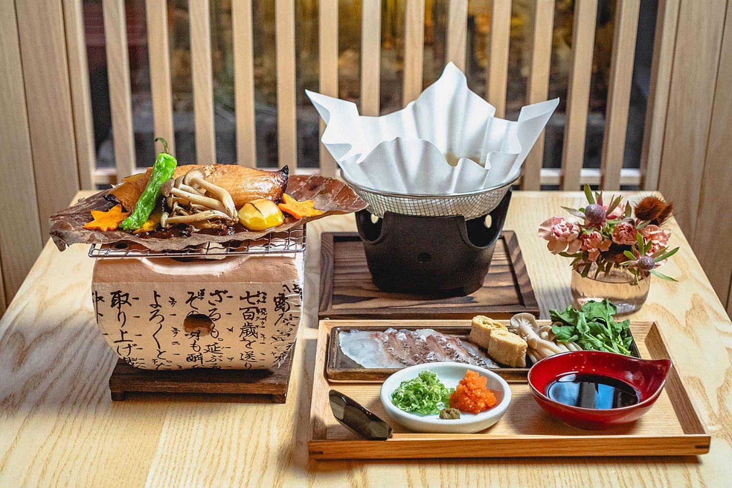 A Japanese meal set on a wooden table with grilled fish and vegetables on a leaf, a hot pot with paper lid, sashimi, dipping sauces, greens, mushrooms, and a small vase of flowers in the background.