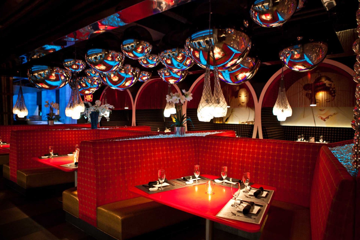 A modern restaurant interior with red upholstered booths, glowing red tables, hanging silver orb lights, and decorative wall art, creating a stylish and colorful atmosphere.