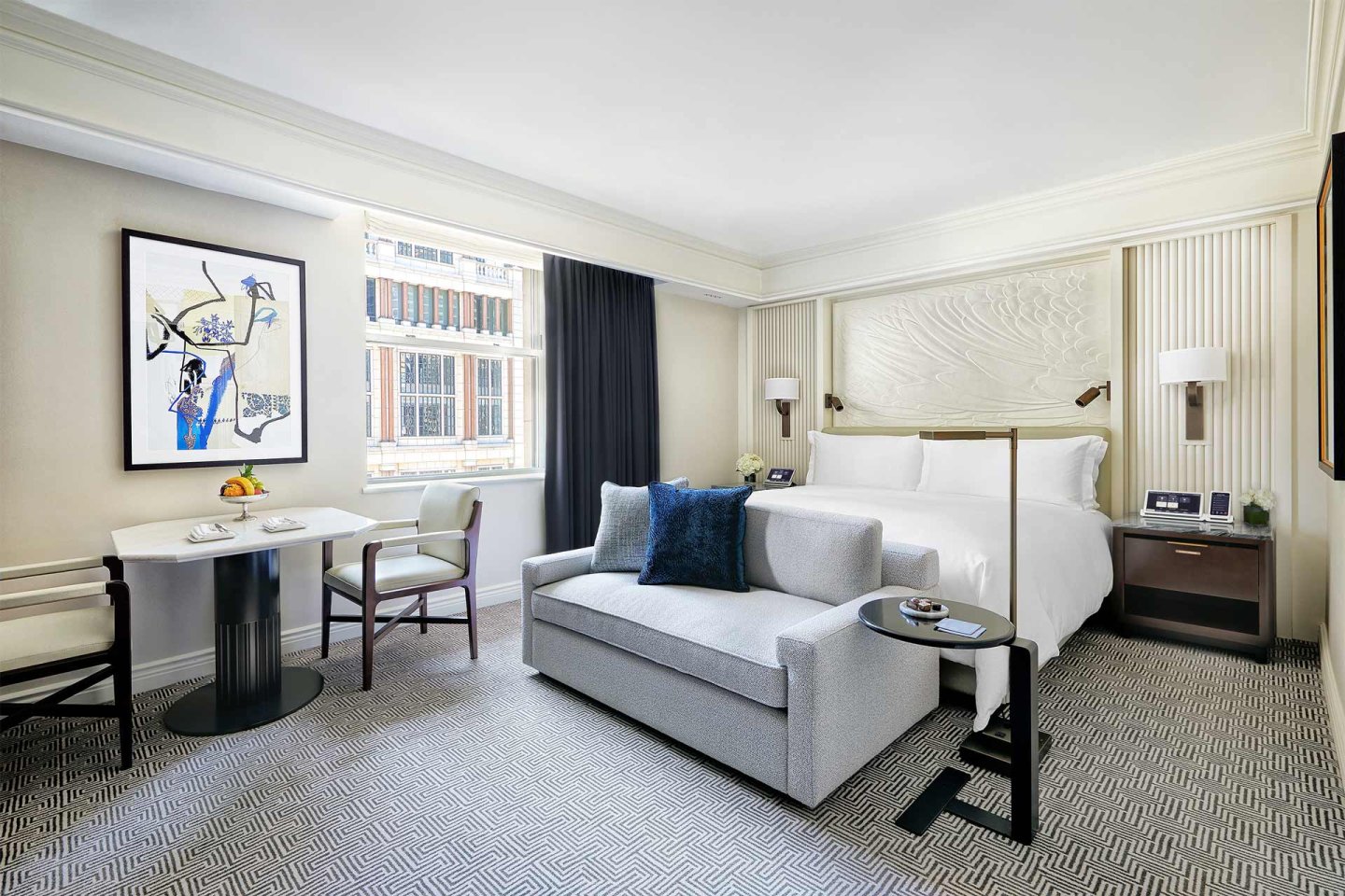 A hotel room with a large bed, light grey sofa, and a dining table with two chairs. A decorative painting hangs on the wall.