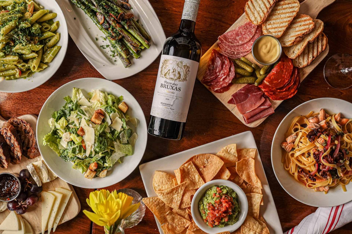 A table set with various dishes, including pasta, salad, grilled asparagus, charcuterie, chips with guacamole, cheese, olives, and a bottle of wine in the center, all arranged on a wooden surface.