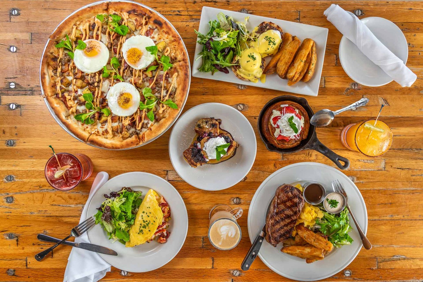 A wooden table set with a variety of brunch dishes, including a breakfast pizza with eggs, steak, eggs Benedict with potatoes, an omelet, salad, toast, coffee, and two colorful juices.