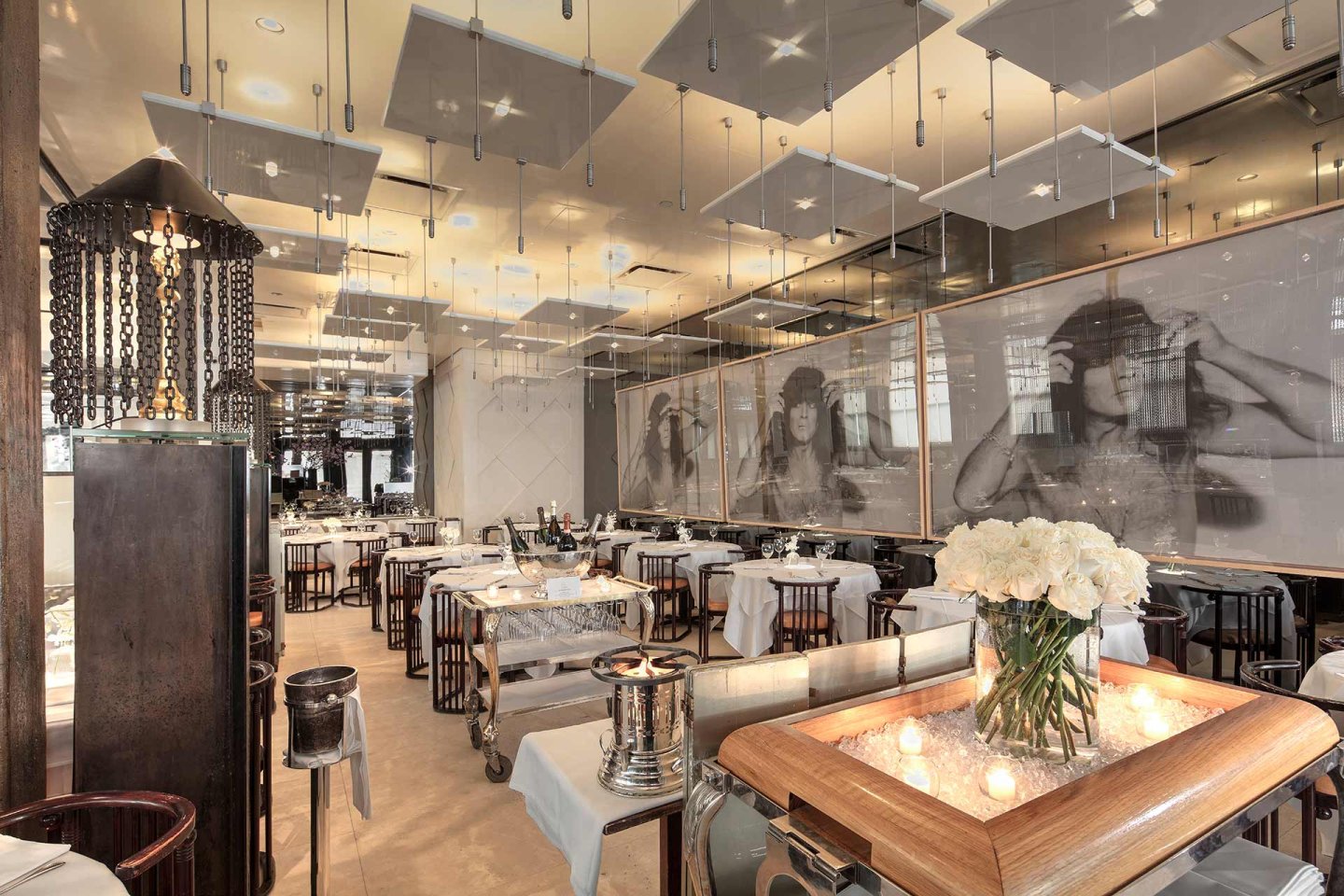 Elegant restaurant interior with neatly set tables, white tablecloths, modern lighting, large black-and-white portraits on the wall, and a centerpiece of white flowers and candles on a wooden cart in the foreground.