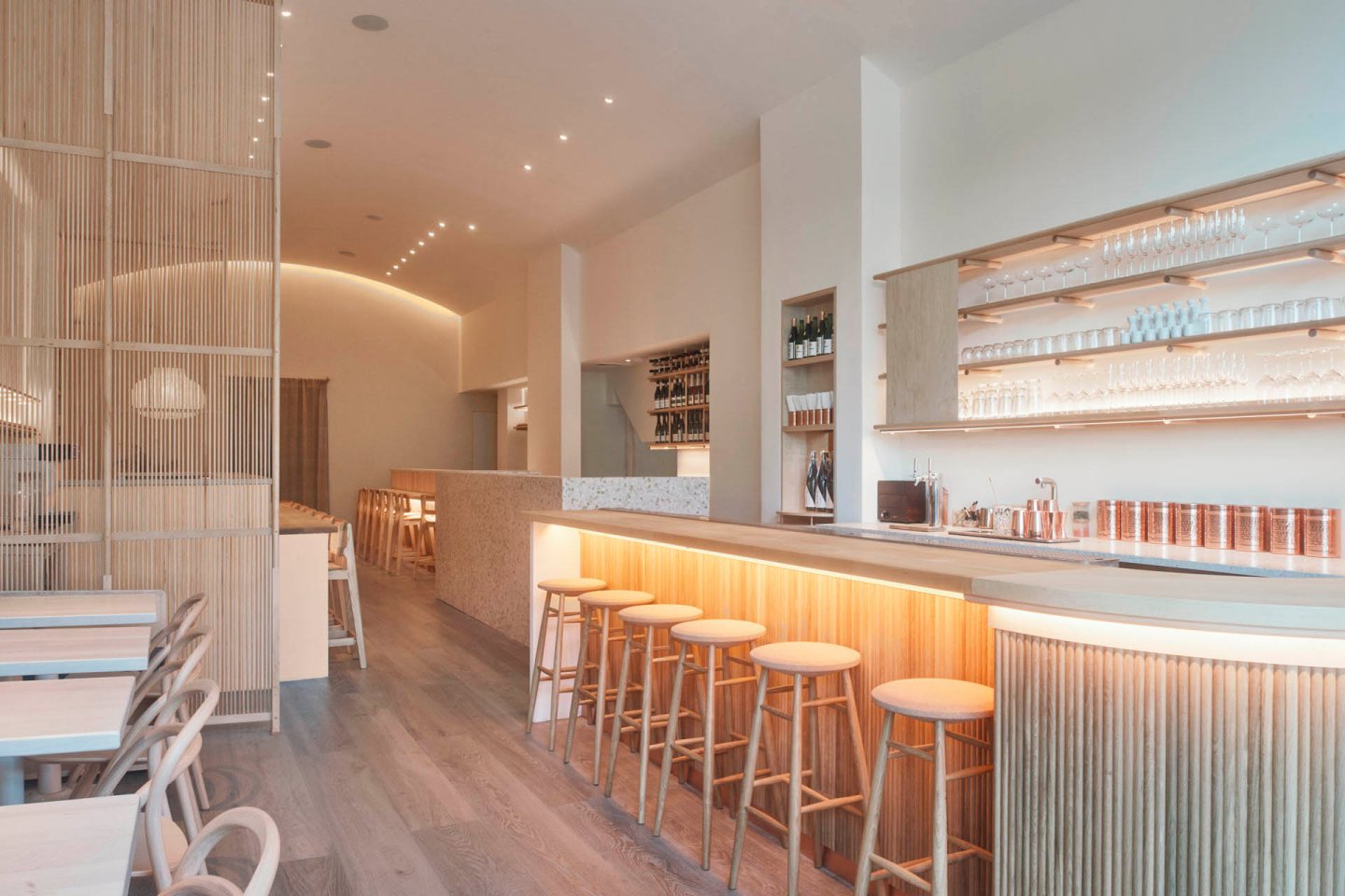 Modern, minimalist interior of a restaurant with light wood accents. Features include a long bar with wooden stools, shelves with glasses, tables and chairs, and subtle overhead lighting creating a warm atmosphere.