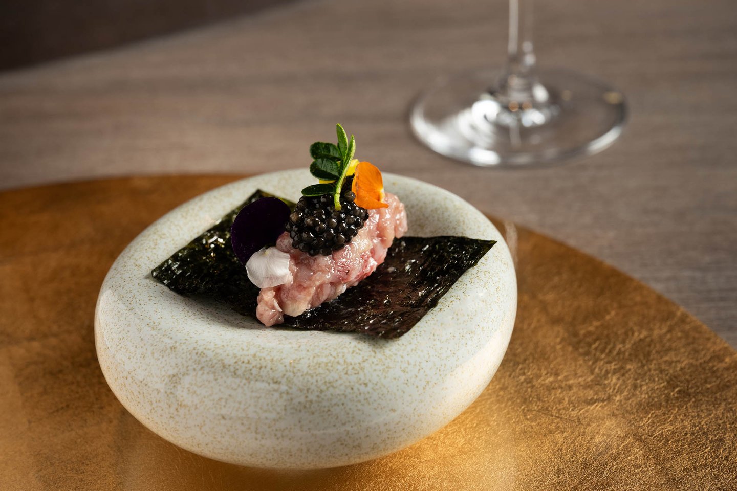 A gourmet dish featuring minced meat topped with caviar, herbs, and edible flowers, served on a sheet of seaweed in a white bowl on a gold surface. A wine glass is blurred in the background.