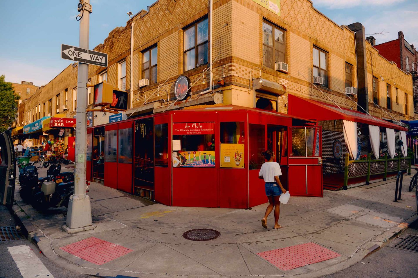 A person walks past a red corner restaurant with outdoor seating on a city street, surrounded by brick buildings, storefronts, and a one-way street sign.