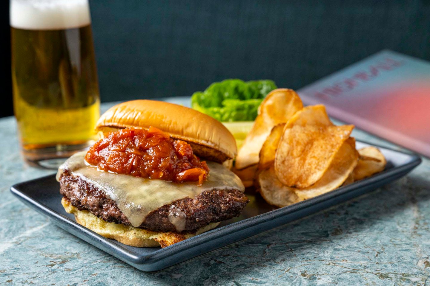 A cheeseburger with sauce sits on a rectangular plate next to a pile of potato chips. A tall glass of beer and a menu are in the background on a marble table.