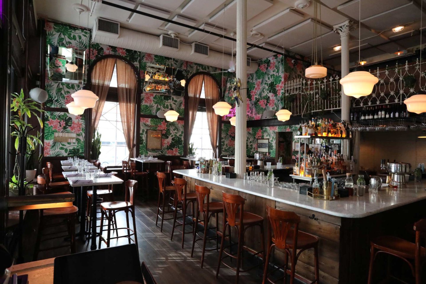A stylish bar with a marble counter, wooden chairs, tropical floral wallpaper, pendant lights, and large windows with sheer brown curtains, creating a cozy and inviting atmosphere.