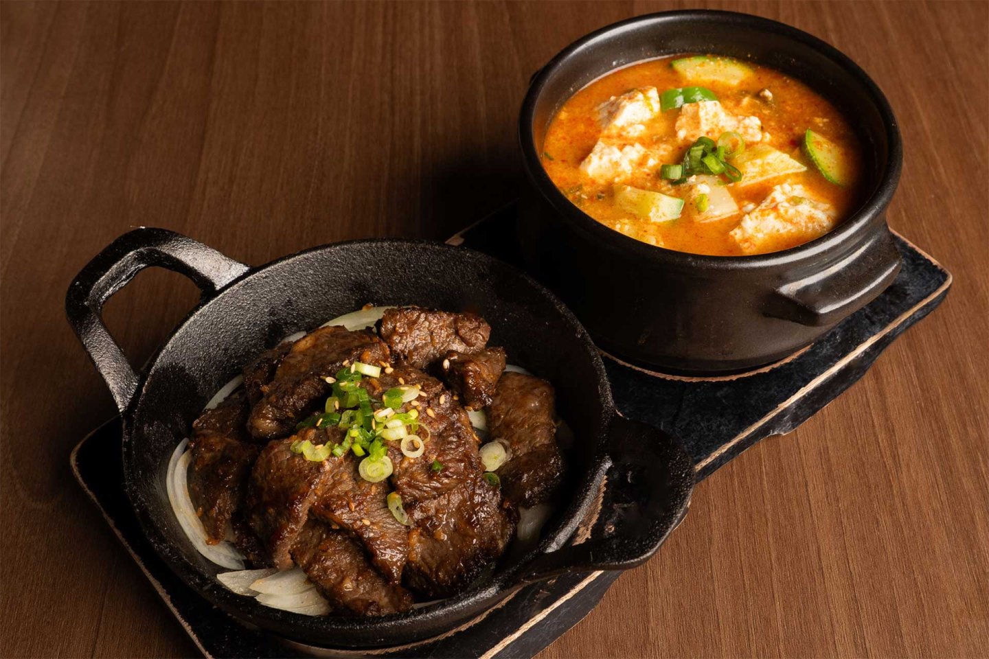 Sliced grilled beef on onions in a cast iron dish next to a black pot of tofu soup with vegetables, both on a dark wooden table.