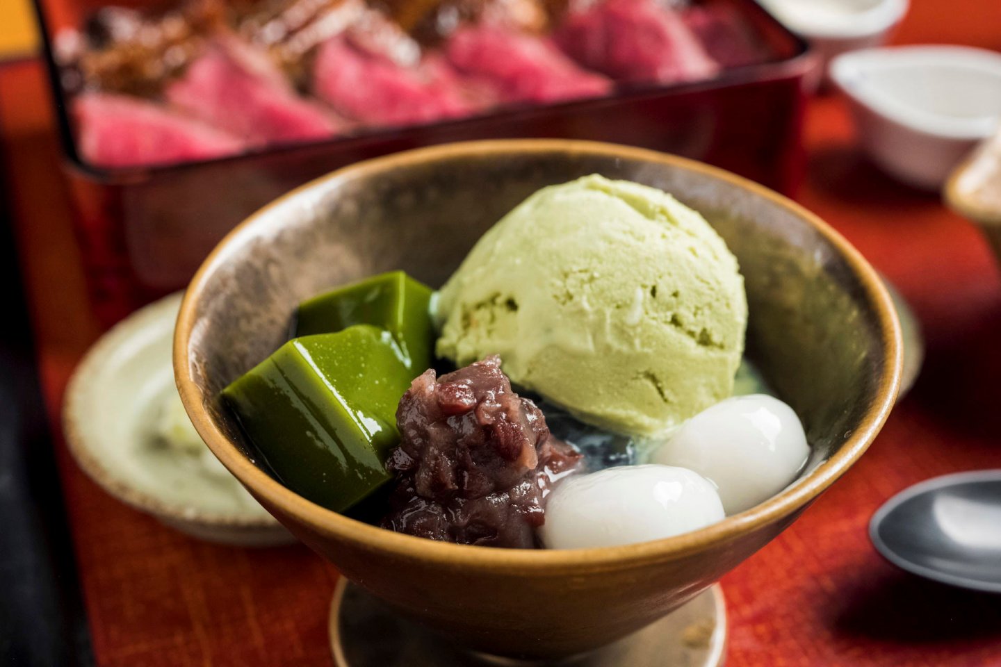 A bowl with green tea ice cream, green jelly cubes, red bean paste, and mochi balls. The background shows a dish of sliced meat, slightly out of focus.