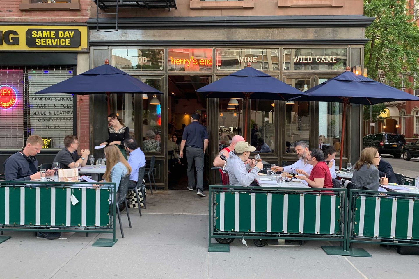 People dine at outdoor tables under navy umbrellas at a city restaurant. The restaurant has large windows and a sign reading "Wine Wild Game." The street has greenery and nearby businesses visible.