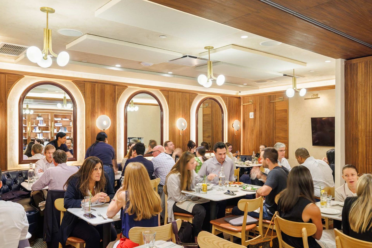 A busy, modern restaurant with wooden walls and round mirrors. Diners are seated at tables eating, drinking, and conversing. Warm lighting and a lively atmosphere fill the space.