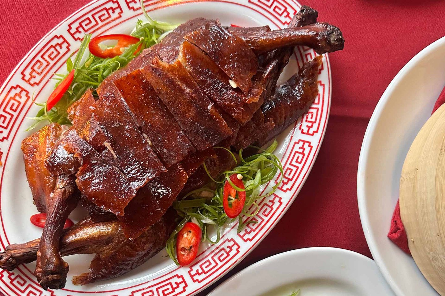 A platter of crispy, sliced Peking duck is served on a decorative plate with red patterns, garnished with sliced green onions and red chili peppers on a red tablecloth.