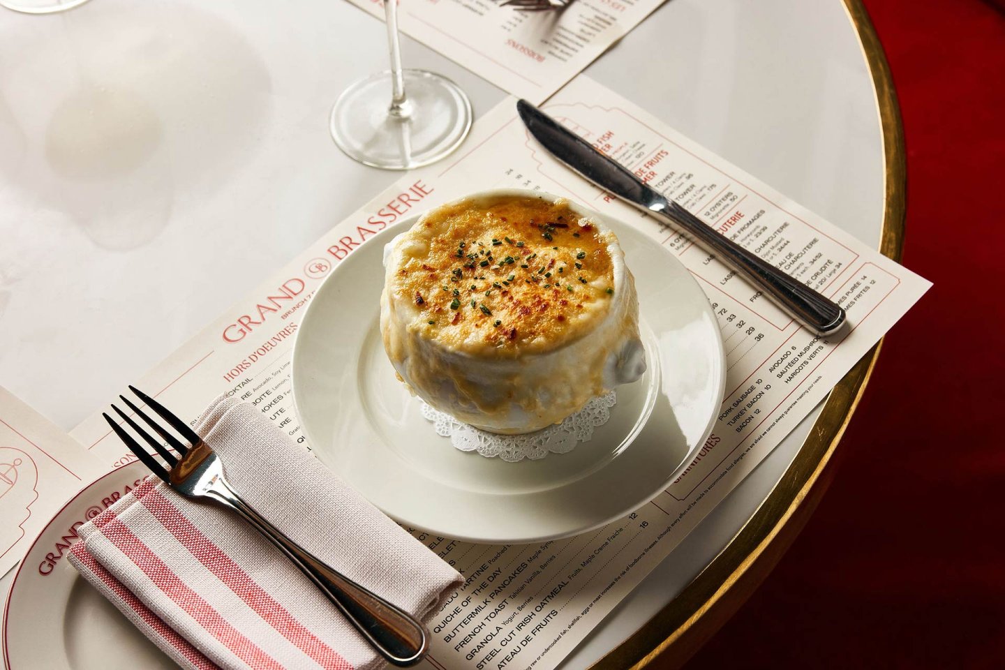 A bowl of French onion soup topped with melted, browned cheese sits on a white plate with a doily, beside a knife and a fork wrapped in a striped napkin on a restaurant table.