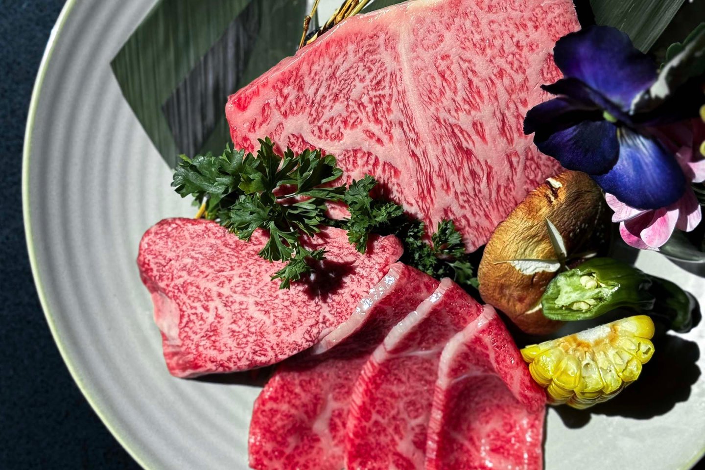 Assorted cuts of marbled raw beef arranged on a plate with parsley, shiitake mushroom, sliced okra, corn, and purple flowers, displayed on a leaf.