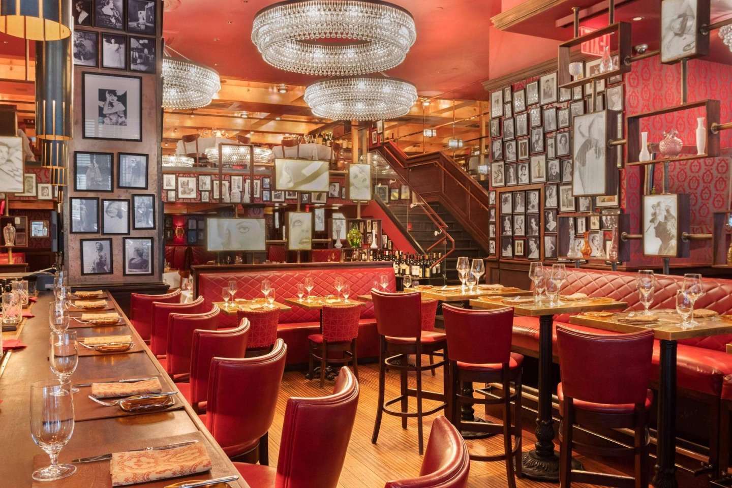 Elegant restaurant interior with red leather chairs, tufted banquette seating, round tables set with glassware, and walls covered in framed black-and-white photos. Two large crystal chandeliers hang from the ceiling.