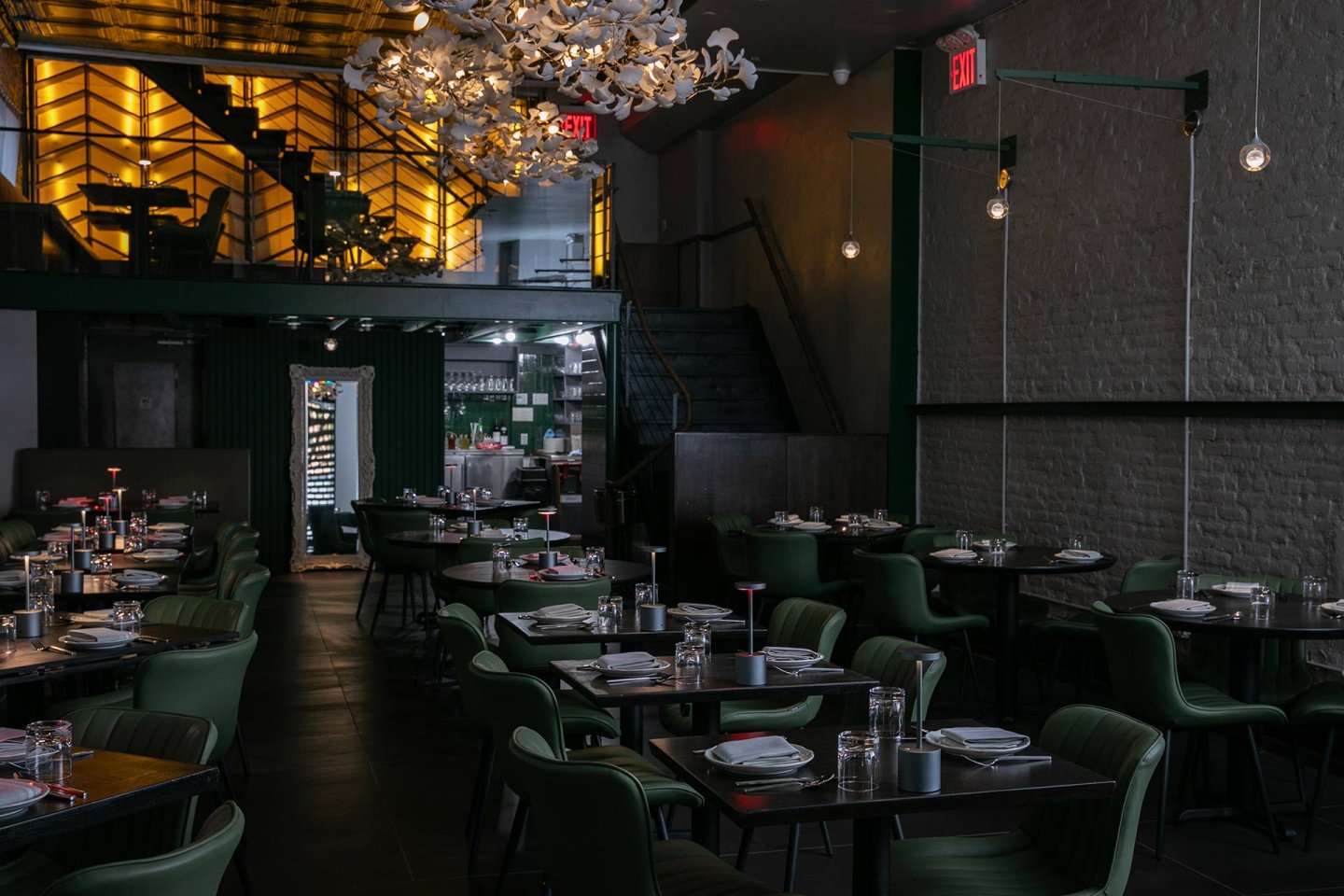 Dimly lit modern restaurant with green chairs, tables set with plates and glasses, a floral chandelier, exposed brick wall, and an illuminated wine rack on a mezzanine level.