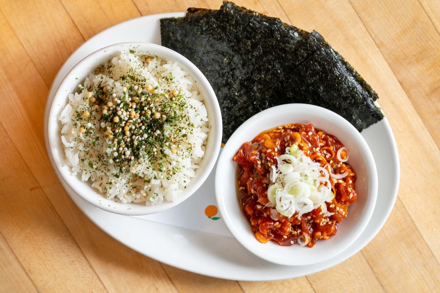 A bowl of white rice topped with seasoning, a bowl of spicy marinated fish garnished with sliced green onions, and several sheets of seaweed on a white oval plate.