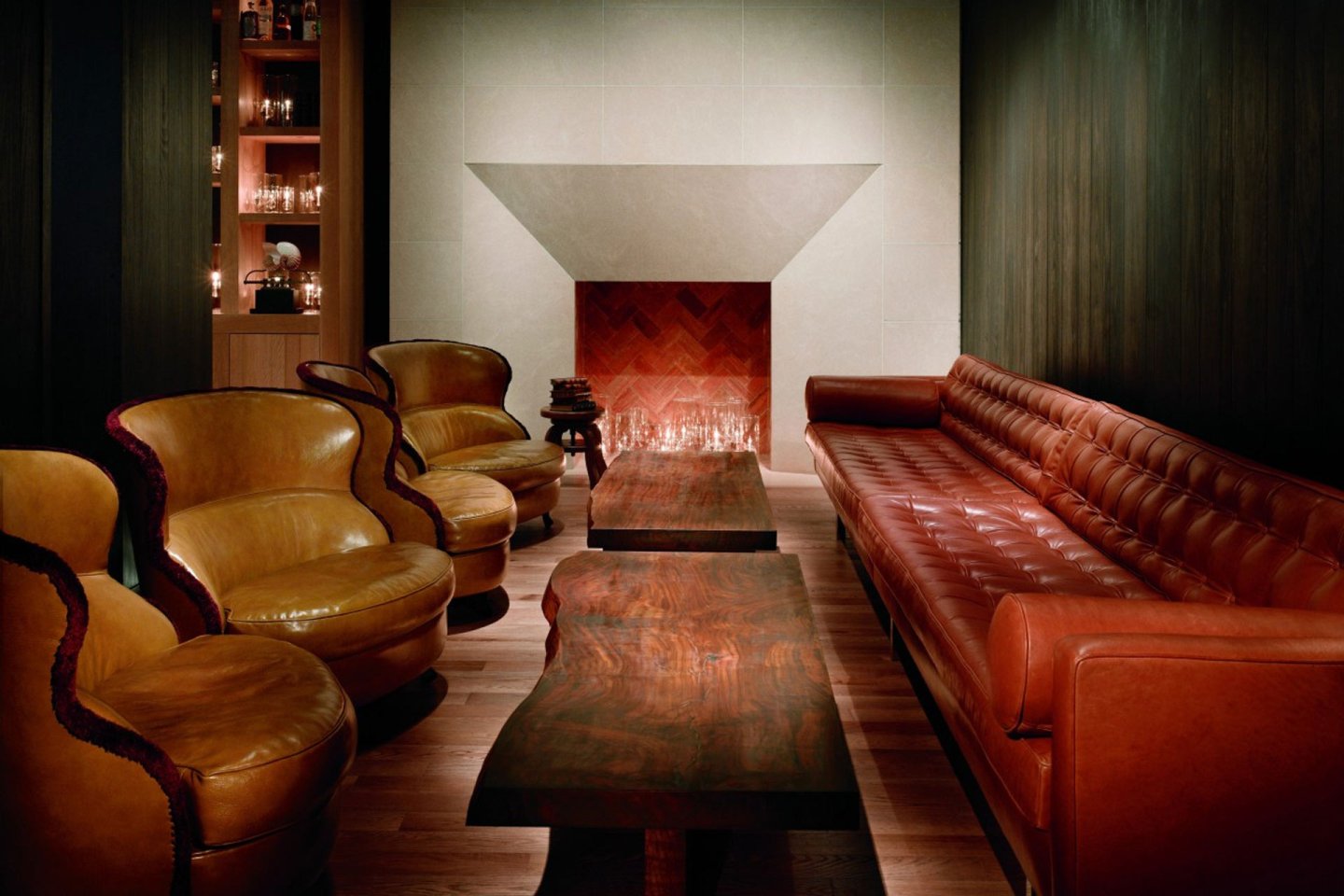 A cozy lounge features two brown leather armchairs and a long brown leather sofa facing each other with two wooden coffee tables in between. A fireplace with stacked logs is in the center, surrounded by dark wood paneling and shelves with glassware.
