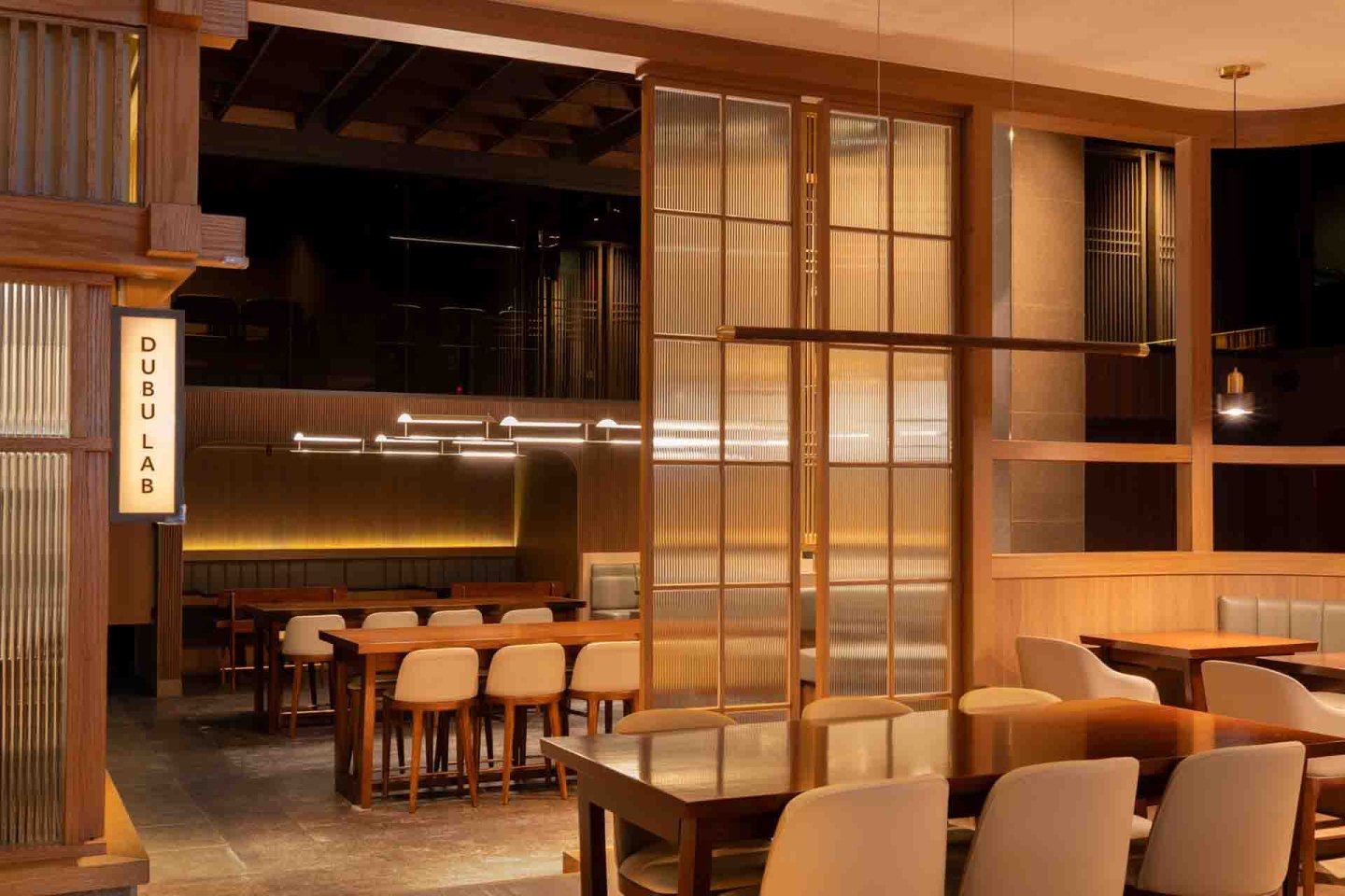Modern restaurant interior with wooden tables and beige chairs, warm lighting, and a sign reading "DUBU LAB" on a wall divider. The space appears clean, organized, and inviting.