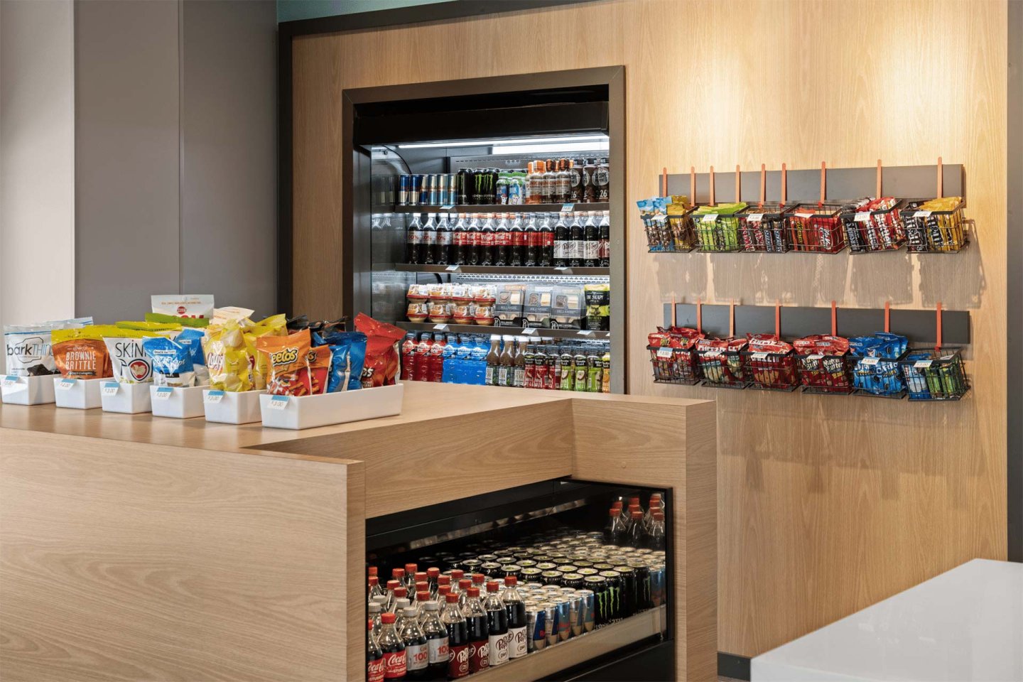 A modern snack bar with shelves and a fridge stocked with assorted drinks, chips, candy, and snacks. Colorful bags and bottles are neatly arranged on wooden counters and wall racks.