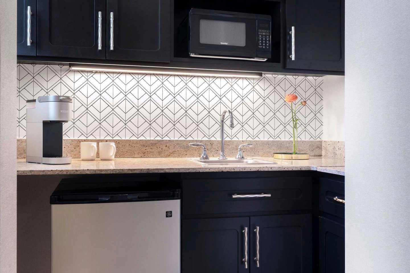 A modern kitchenette with black cabinets, a small refrigerator, a microwave, a coffee maker with two mugs, a sink, and a single flower in a vase on the counter, set against a geometric tiled backsplash.