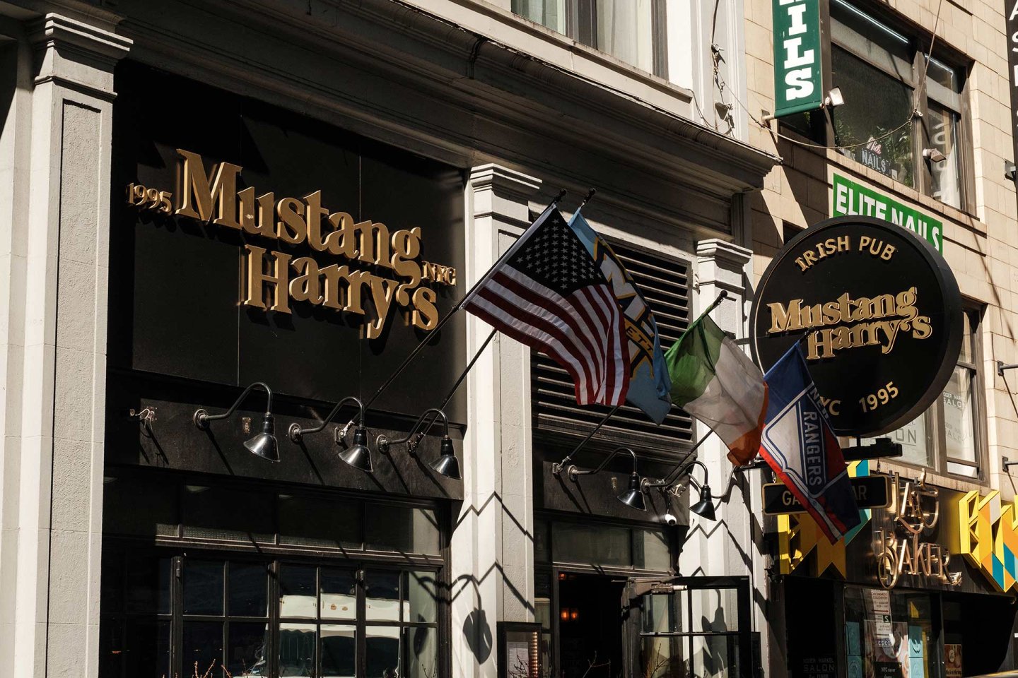 Exterior of Mustang Harry’s Irish pub, with its sign and American, Irish, and New York City flags displayed above the entrance. Sunlight casts shadows on the building’s facade. Signs for “Elite Nails” are visible nearby.