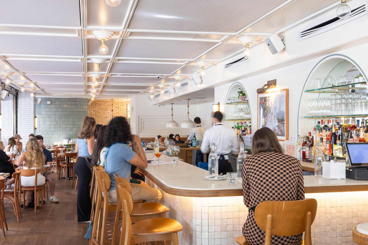 A bright, modern restaurant with wooden chairs and tables, a curved bar, and people dining and chatting. Bartenders work behind the bar, and shelves of glasses and bottles are visible.
