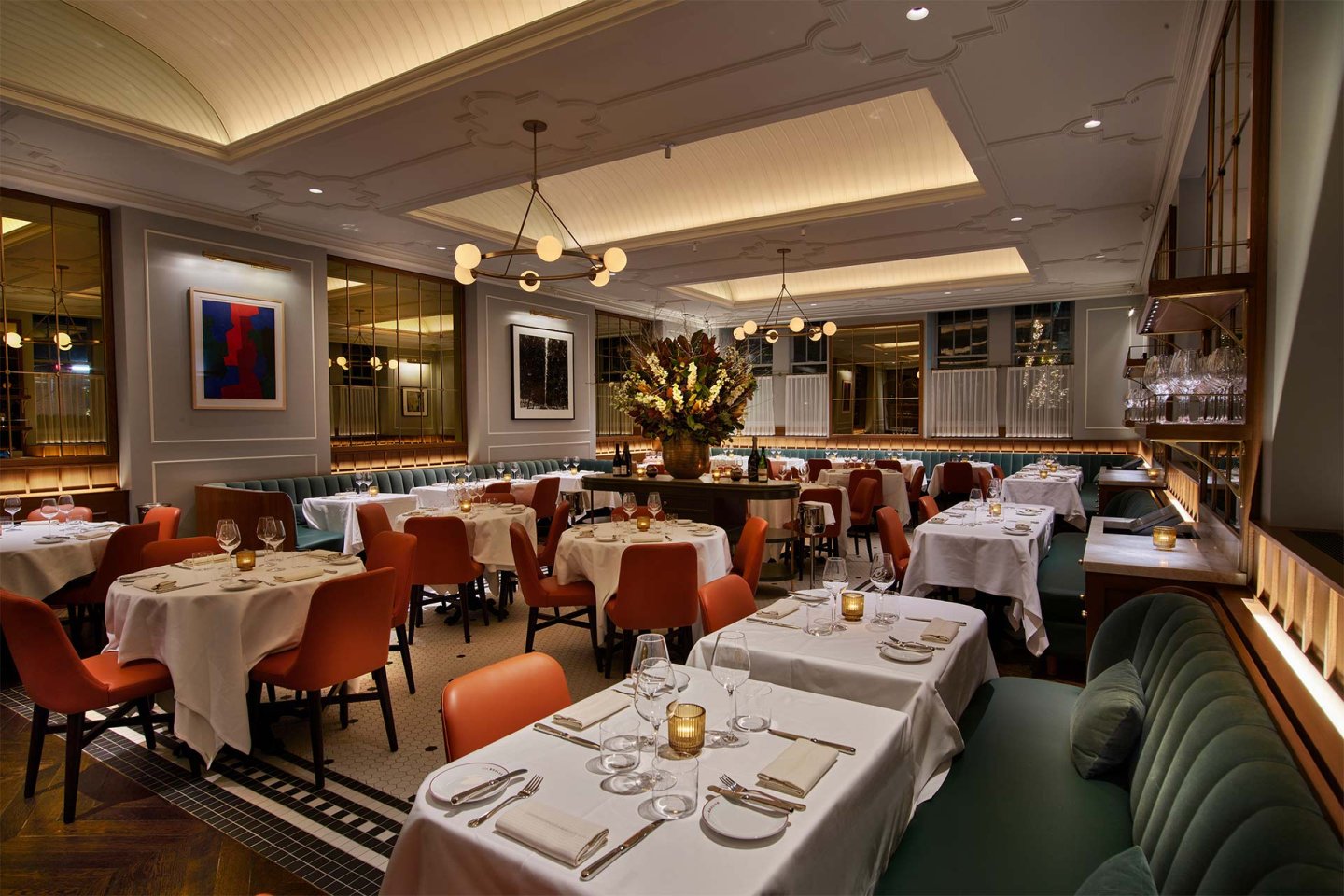 Elegant restaurant interior with neatly set tables, orange chairs, green banquettes, soft lighting, modern art on the walls, and a large floral centerpiece creating a warm, inviting atmosphere.