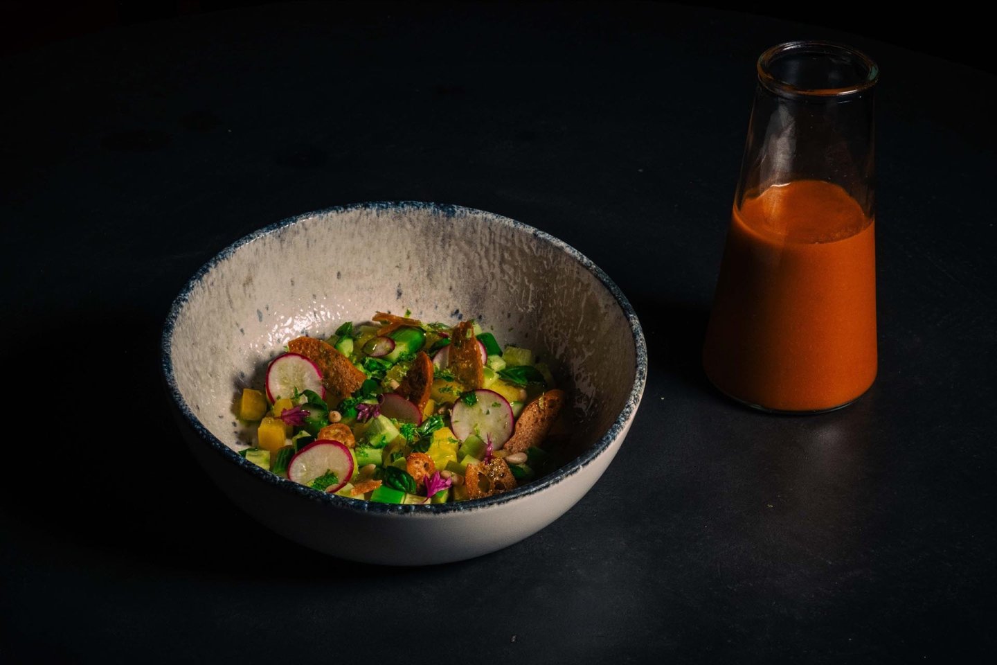 A ceramic bowl with a colorful salad of radishes, greens, and croutons sits on a dark surface next to a glass bottle filled with orange dressing.