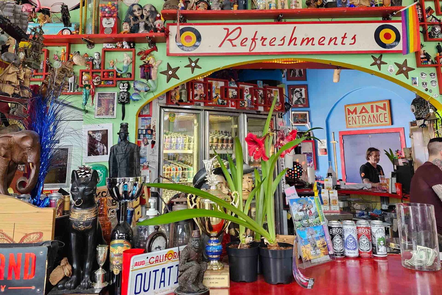 Colorful refreshment stand with plants, trophies, drinks in coolers, quirky decor, and a "Main Entrance" sign. Two people are behind the counter, one taking an order.