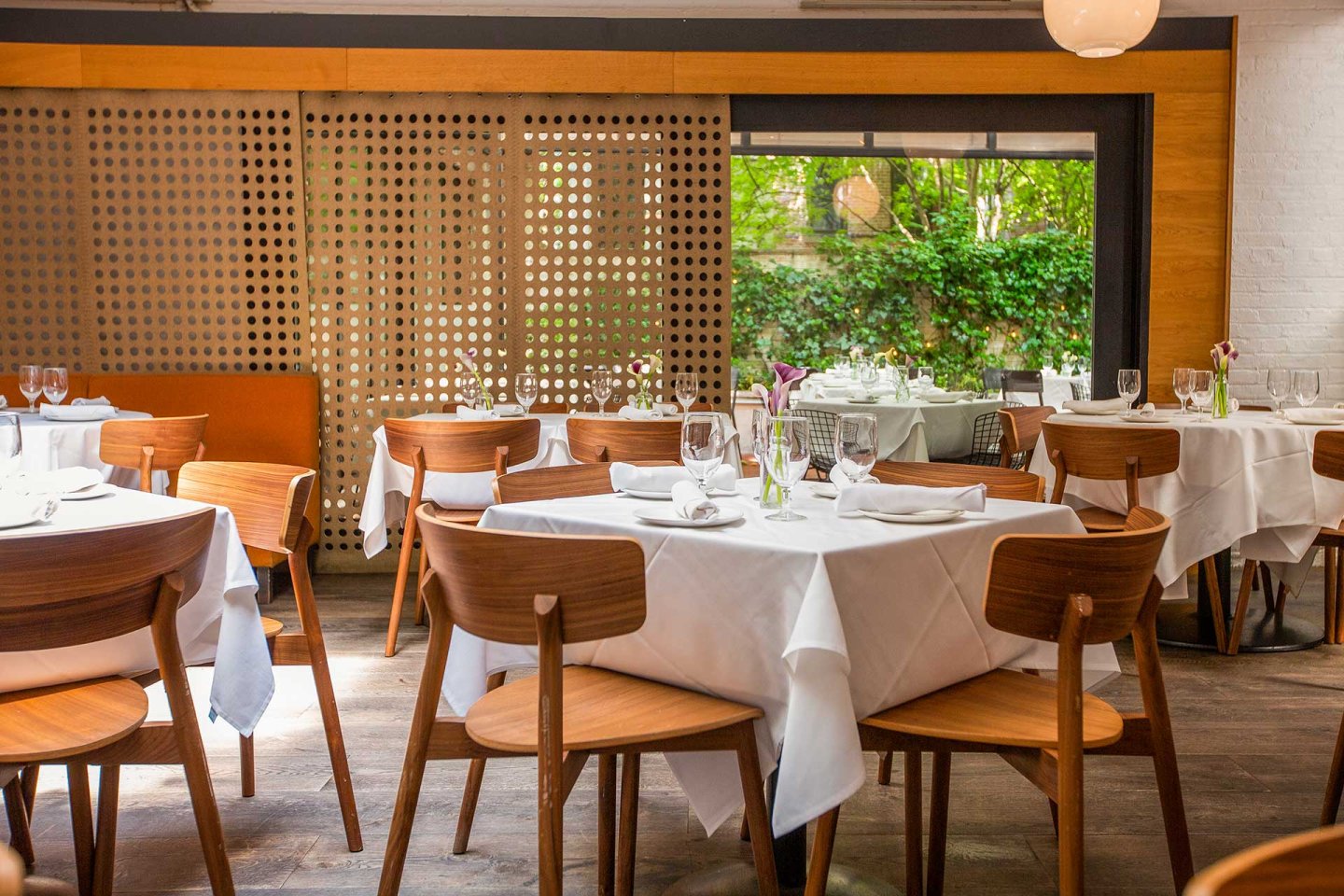 A bright, modern restaurant dining area with wooden chairs and tables covered in white tablecloths, set with glassware and folded napkins. Large window shows greenery outside; perforated wood panel decorates the wall.