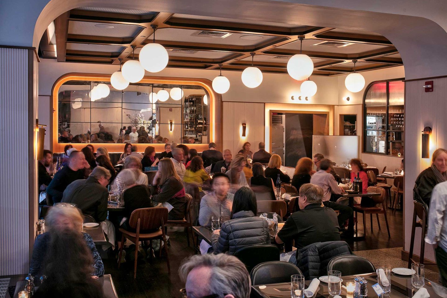 A stylish, modern restaurant filled with people dining at tables. Warm lighting from globe lamps and wall sconces creates a cozy atmosphere. A large mirror reflects the lively crowd and decor.