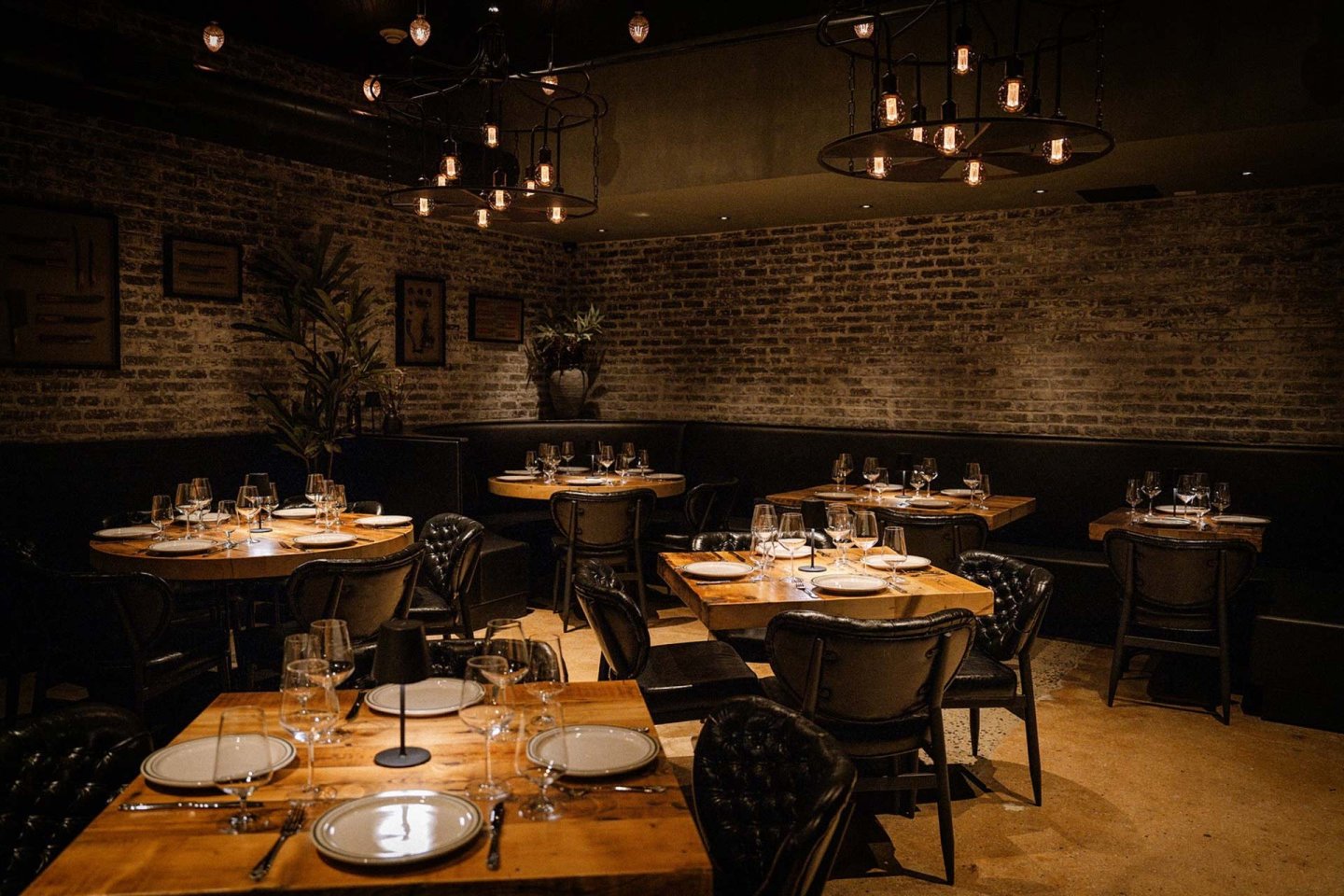 A cozy, dimly lit restaurant with exposed brick walls, wooden tables set with plates, cutlery, and glasses, black cushioned chairs, decorative plants, and hanging industrial-style light fixtures.