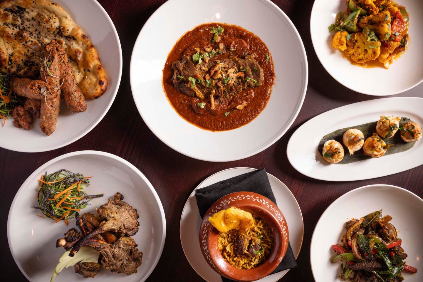 A variety of Indian dishes arranged on a dark wooden table, including curry, naan, biryani, stuffed vegetables, and grilled items. Each dish is presented in white bowls or plates, garnished with herbs and colorful vegetables.