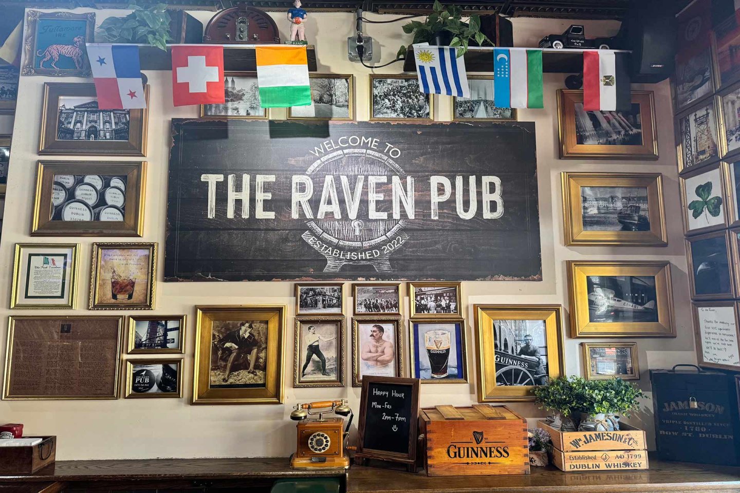 Wall of The Raven Pub decorated with framed photos, memorabilia, shelves, and small flags. A large sign reads "The Raven Pub." Guinness and Jameson boxes are on a counter beneath the sign.
