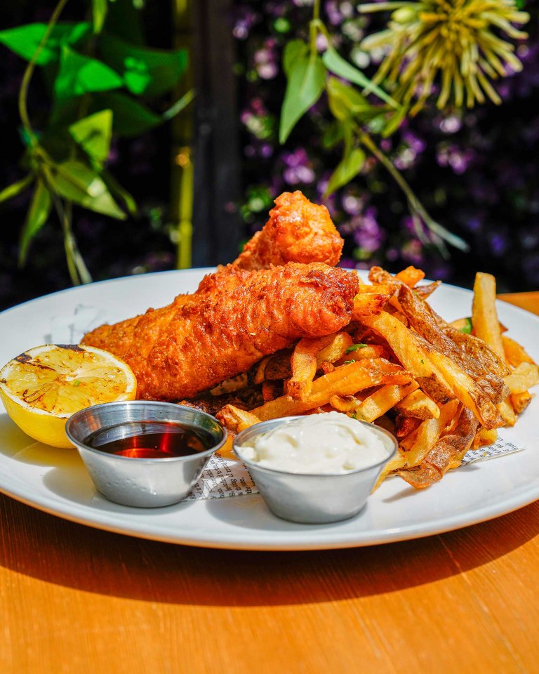 A plate of crispy battered fish and golden fries served with a charred lemon half, a cup of tartar sauce, and a cup of dark dipping sauce, set on a wooden table with a leafy, flowery background.