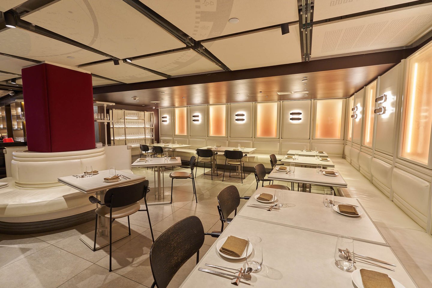 Modern restaurant interior with beige and brown tones, white tables set with plates and napkins, cushioned seating along the wall, soft lighting, and a clean, minimalist design. Room appears empty and ready for guests.