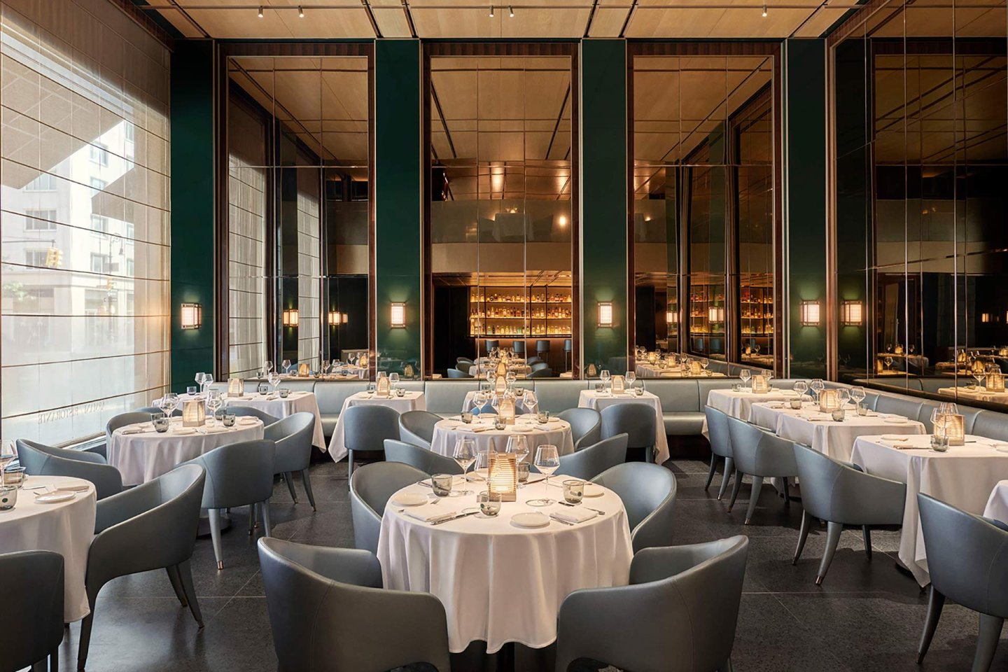 Elegant restaurant interior with round tables covered in white tablecloths, surrounded by modern grey chairs; soft lighting, mirrored walls, and a stylish bar area in the background create a sophisticated atmosphere.