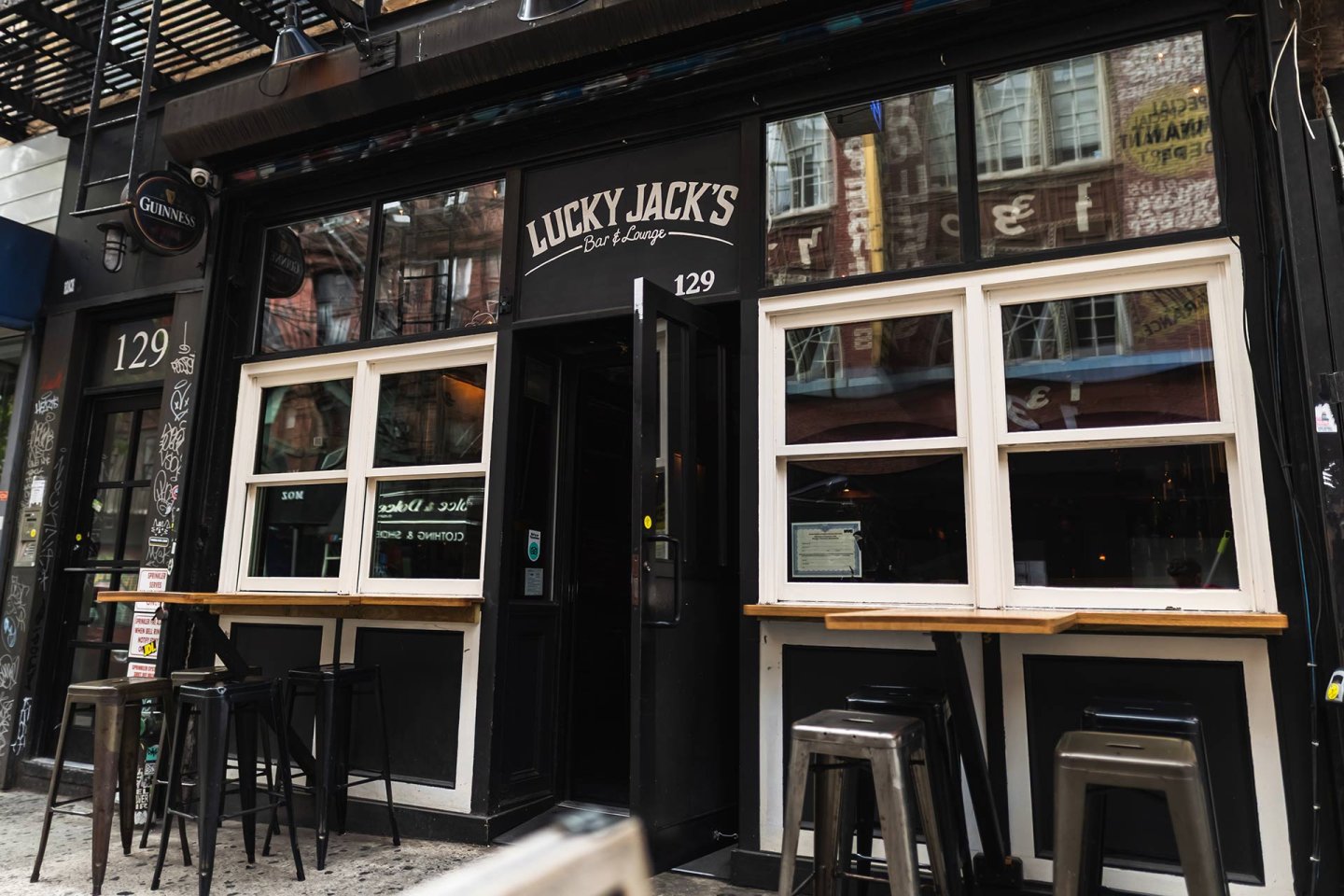 Street view of Lucky Jack’s Bar & Lounge with large windows, bar stools, a black exterior, and address number 129 displayed above the entrance. Reflections of nearby buildings are visible in the windows.