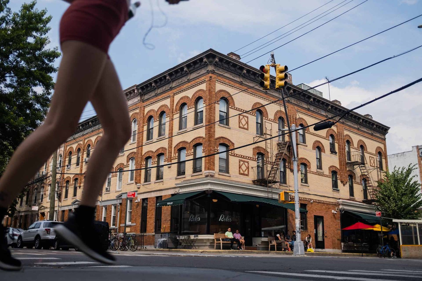 A person in athletic clothing jogs across a city street in front of a historic brick building with shops and a café at the corner, under a partly cloudy sky.