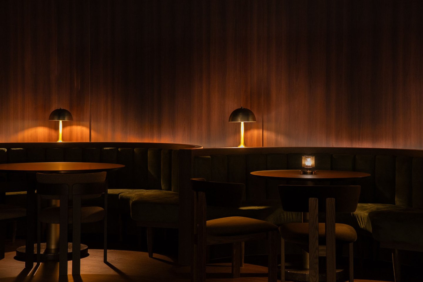 Dimly lit lounge with round wooden tables, curved velvet banquettes, and warm light from modern table lamps against a dark wooden wall.