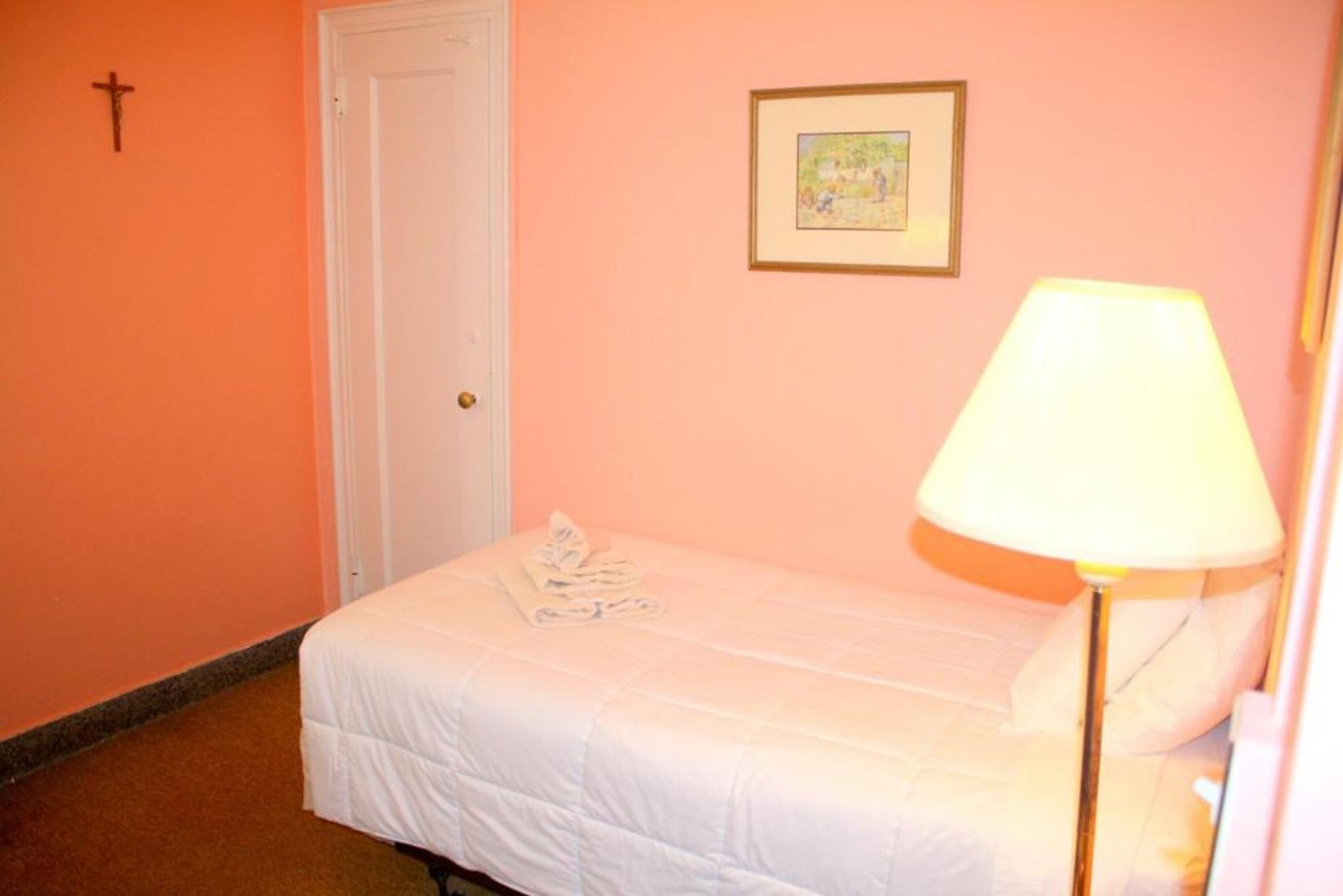 A small bedroom with peach-colored walls, a single bed with a white comforter, a towel folded on the bed, a framed picture and a crucifix on the wall, and a lit table lamp beside the bed.