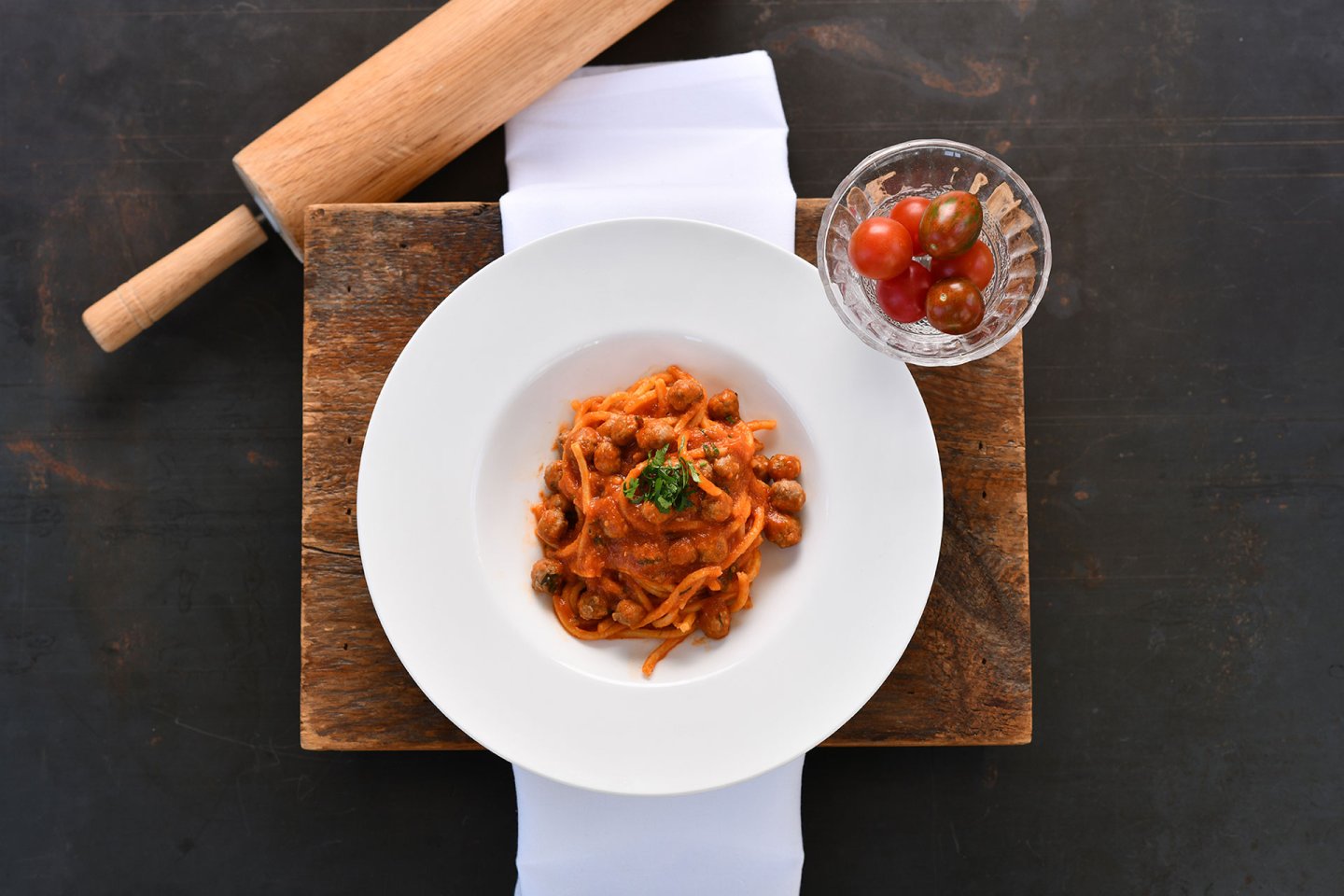 A white plate of pasta with tomato sauce is garnished with herbs, placed on a rustic wooden board with a white napkin. A glass bowl of cherry tomatoes and a wooden rolling pin are nearby.