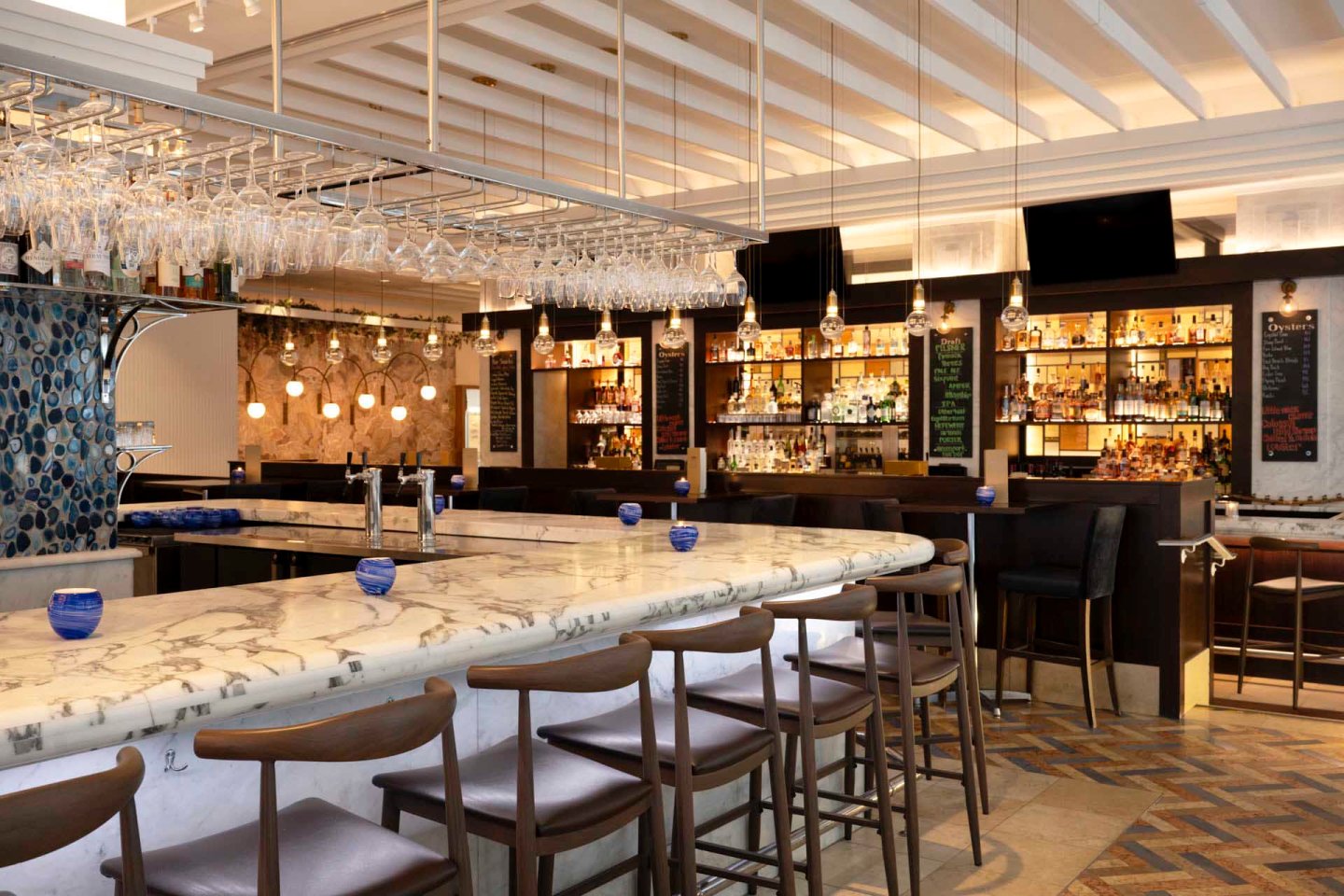 A stylish bar with a marble countertop and wooden barstools. Shelves stocked with bottles are visible in the background. Hanging wine glasses and pendant lights add a touch of elegance. The floor features a geometric pattern.